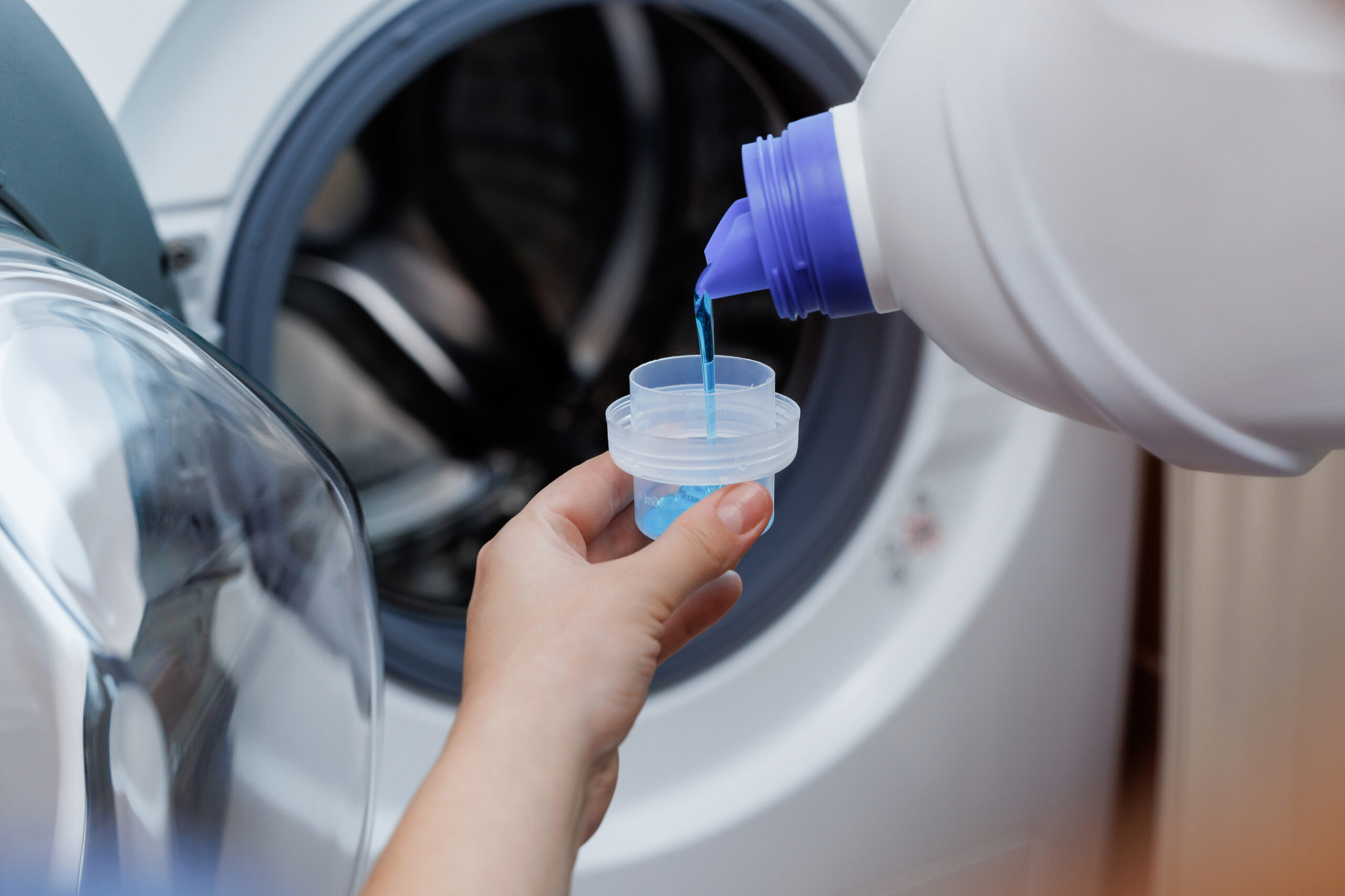 Close up shot of a woman hands squeezing liquid gel laundry detergent from a plastic bottle into a small container, with a washing machine in the background